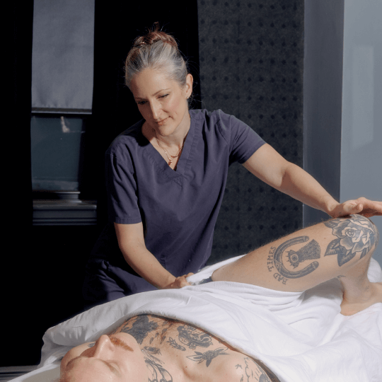 A massage therapist giving a massage to a patient who is face up and having his leg stretched. She is in blue scrubs and he is in white sheets