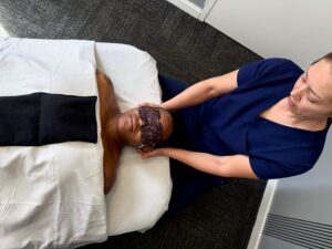 Massage therapist in blue scrubs giving a neck massage to someone under a weighted blanket. 